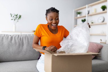 Karton paketleri açan mutlu siyahi kadın, başarılı alışverişe duygusal tepki veriyor, plastik ambalajı çıkarıyor, evden büyük alışverişlerle tatmin oluyor. Çevrimiçi mağaza ve dağıtım kavramı
