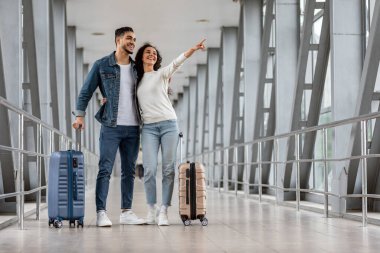 Seyahat Reklamları. Havalimanında dikilen ve yan yana duran mutlu genç Orta Doğulu çift, bavulları olan, terminalde bekleyen, birlikte seyahat etmekten keyif alan neşeli Arap eşler.