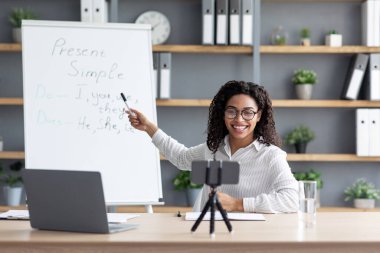 Covid-19 salgını, yeni normal, sanal sınıf, ders ve eğitim, evde eğitim. Milenyum güzeli, siyah gözlüklü kadın öğretmen kara tahtayı gösteriyor. Oturma odasındaki telefon kamerasına bakın.
