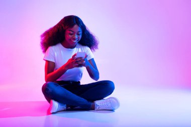 Genç bir Afrikalı Amerikalı kadın bacak bacak üstüne atmış bir şekilde oturuyor, cep telefonunu bir arkadaşıyla sohbet etmek için kullanıyor. Etrafı yumuşak neon ışıklarıyla çevrili. Sosyal medyada canlı bir atmosfer yaratıyor..