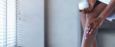 Bir kadın iyi aydınlatılmış bir odada oturuyor ve bacağına krem sürüyor. Bir elinde küçük bir krema kavanozu tutuyor. Güneş ışığı pencerenin perdelerinden süzülüp duvarda desenler oluşturur..