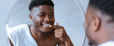 Bir adam banyoda dikilip aynada kendine bakıyor. Gülümsüyor ve dişlerini gösteriyor. O, omuzlarına bir havlu takar. Ayarların yumuşak ışıklandırması ve basit dekorasyonu var.