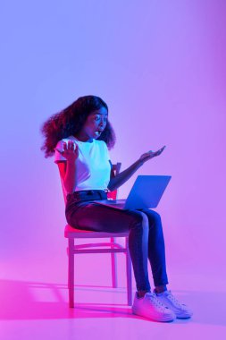 Neon ışıklı bir yerde dizüstü bilgisayarıyla bir sandalyeye oturan siyahi bir kadın şaşırdı. İfadesi, çevrimiçi çalışması sırasında yaptığı bir hataya şaşırdığını gösteriyor..