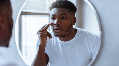 Yuvarlak aynanın önünde duran bir adam yansımasına bakıyor. Görünüşüne konsantre olurken yüzüne dokunuyor. Yakınlardaki pencerelerden doğal ışık giriyor.