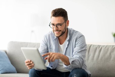 Boş zaman kavramı. Dijital tablet kullanan, oturma odasındaki koltukta oturan, fotokopi çeken mutlu bir adam. Neşeli adam sosyal medyayı kontrol ediyor ya da arkadaşlarıyla sohbet ediyor.