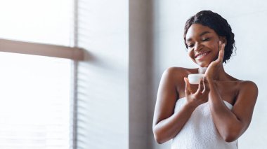 Elinde küçük bir cilt ürünüyle pencerenin yanında duran bir kadın. Yüzüne nazikçe dokunurken gülümsüyor, bir anlık özgüven gösterisi sergiliyor. Doğal ışık odayı aydınlatır..