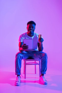 Neşeli siyah bir adam elinde dizüstü bilgisayar ve kredi kartı olan bir sandalyede oturuyor ve online alışverişin tadını çıkarıyor. Parlak neon lambaları ödemesini yaparken canlı bir atmosfer yaratıyor..