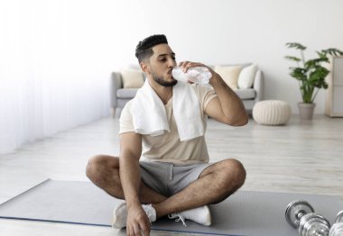 Yoga minderinde bağdaş kurmuş, ev içi eğitimden sonra su içen yakışıklı genç bir Arap. Çekici bir orta çağ Orta Doğulu adam ev çalışması sırasında susuz kalıyor.