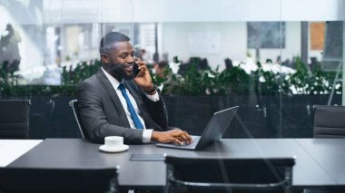 Resmi giyinmiş bir adam ofisteki bir konferans masasında oturuyor. O bir dizüstü bilgisayar kullanıyor ve gülümserken telefonla konuşuyor. Bitkiler arka planda görünür..