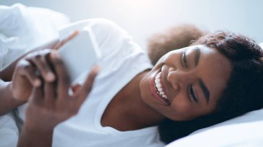 Bir kişi bir yatakta uzanmış, akıllı telefona bakarken gülümsüyor. Aydınlatıcı ve rahat bir ortam var..