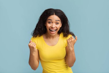 Evet, harika haberlerim var. Mavi stüdyo arka planında izole edilmiş, çığlık atan ve titreyen siyahi kadın portresi. Neşeli Afrikalı Amerikalı kadın tezahürat yapıyor ve bağırıyor, zaferi kutluyor.