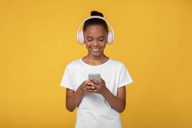 Beyaz tişörtlü, kulaklıklı, telefonda video izleyen, sarı arka planda izole edilmiş, boş bir alanda gülümseyen genç siyah kız. Çevrimiçi ders, aygıtla çalışma, oyun oynama, sosyal ağlarda sohbet