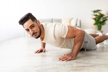 Attractive young Arab man standing in plank pose or doing push ups on mat at home. Fit middle Eastern guy leading active lifestyle, exercising indoors during coronavirus lockdown