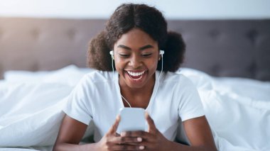 Genç bir kadın beyaz çarşaflar ve koyu bir yatak başlığıyla yatakta oturuyor. Kulaklık takıyor ve akıllı telefonuna gülümsüyor. Sabah saatlerinin keyfini çıkarıyor..