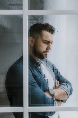 Derin depresyon, sosyal temastan kaçınma, evde kendini soyutlama. Endişe ve agorafobi. Çapraz kollu üzgün, yakışıklı sakallı bin yıllık adam pencereye bakar, ıslak camdan bakar, boşluğu taklit eder.