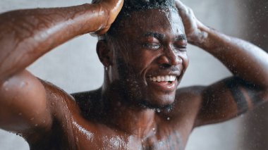 Bir adam duşta akan suyun altında duruyor. Yüzüne ve vücuduna su damlarken çok fazla gülümsüyor. Ayarlar yumuşak, doğal ışığın içeri girdiği bir banyo..