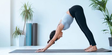 Bir kadın parlak bir odada paspasın üzerinde aşağı doğru köpek yogası yapıyor. Yeşil bitkiler görülebilir, kapalı alandaki atmosfere eklenir. Ortam temiz ve ferah, egzersiz için ideal..