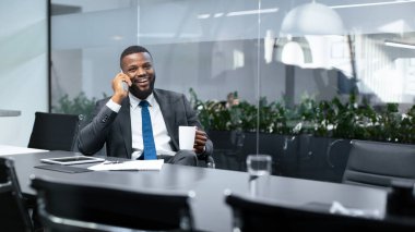 Ofiste oturan bir adam telefonda konuşuyor. O gülümsüyor ve bir kahve fincanı tutuyor. Masada belgeler ve bir tablet var. Bitkiler arka planda görülebilir.