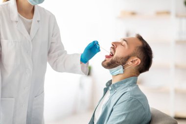 Kadın doktor klinikteki genç adam için oral koronavirüs PCR testi yapıyor. Millennial adam covid tanı prosedüründen geçiyor. Bulaşıcı hastalıkların önlenmesi ve korunma kavramı
