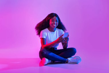 Genç bir Afrikalı Amerikalı kadın internette sörf yapmak ve sohbet etmek için cep telefonunu kullanıyor. Neon ışıkları, dijital bağlantısının keyfini çıkarırken canlı bir atmosfer yaratıyor..