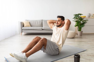 Spor yapan genç bir Arap, karın kası egzersizi yapıyor, evde çalışıyor, boy boy portresi yapıyor. Atletik Orta Doğulu adam covid salgını sırasında aile içi eğitim yapıyor, çekirdek kaslarını güçlendiriyor.