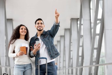 Pasaportları ve biletleri olan genç bir Orta Doğulu çift havaalanında dikiliyor ve birlikte seyahat eden mutlu bir Arap erkek ve kadın Terminal 'deki uçuş kapısını kontrol ediyor,