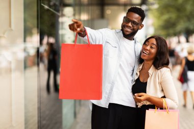 Ticari mal. Gülümseyen Siyah Aile, Alışveriş Torbası taşıyan, Alışveriş Merkezi 'nin önünden geçen, Erkek Müşteri Market Vitrininde Parmak Gösteren, Ürün Ekranı gösteren. Mevsimlik Satış, Büyük Teklif