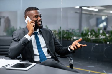 Bir işadamı ofisteki bir masada oturur. Telefonda konuşuyor ve gülümsüyor. Kağıt ve kalem masanın üstünde. Kapalı alan bitkileri arka planda görünür..