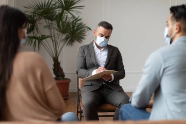 Ofiste oturan psikoterapi seansı sırasında maske ve takım elbise giyen profesyonel erkek psikolog not alıyor. Randevu sırasında aile terapisti ile konuşan genç çift.