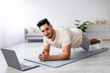 Çevrimiçi sporlar, tecrit sırasında. Evde laptopun önünde egzersiz yapan güçlü genç bir Arap. Doğulu formda bir adam yoga minderinde çalışıyor, dirsek tahtasında duruyor, video dersini takip ediyor.