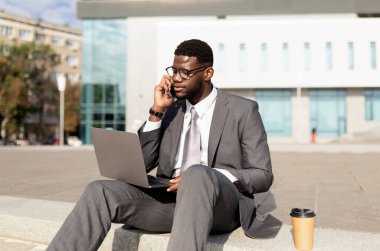 Dışarıda dizüstü bilgisayarı olan siyahi bir işadamı oturuyor ve akıllı telefondan konuşuyor, arka planda ofis binası, kopyalama alanı. Çevrimiçi çalışan kendine güvenen girişimci