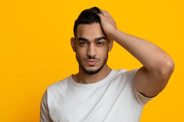 Çekici, sakallı, beyaz tişörtlü ortadoğu çocuğu sarı stüdyo arka planında poz verirken saçına dokunuyor, bayanlarla flört ediyor, yakın plan portresi, fotokopi uzayı kopyalıyor.
