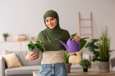 Kafiyeli genç bir Arap kadın, elinde bir su kovası ve kapalı bir saksı bitkisi tutarken gülümsüyor. Oturma odasında canlı bitkilerle sıcak ve davetkar bir atmosfer yaratıyor..