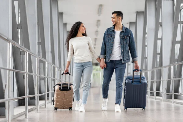 Mutlu Gezginler. Havaalanında valizlerle yürüyen genç Arap çiftin portresi, bavullarını taşıyan ve ayrılış kapısına giderken birbirlerine gülümseyen romantik ortadoğulu eşler,