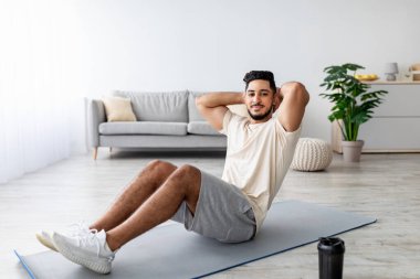 Atletik bin yıllık Arap adam karın egzersizi yapıyor, evde çalışıyor, tam boy. Koronavirüs salgını sırasında ev içi antrenman yapan ve iç kaslarını güçlendiren formda bir Doğulu.