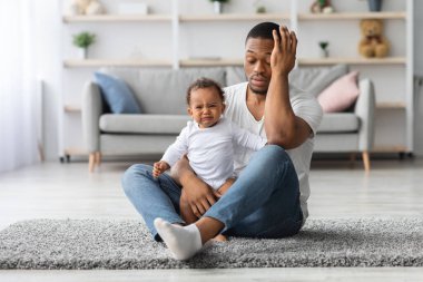 Yorgun, genç bir baba evde yerde ağlayan yeni doğmuş bebekle oturuyor, umutsuz Afrikalı Amerikalı baba bebeğini üzüyor, babalık izni sırasında acı çekiyor.