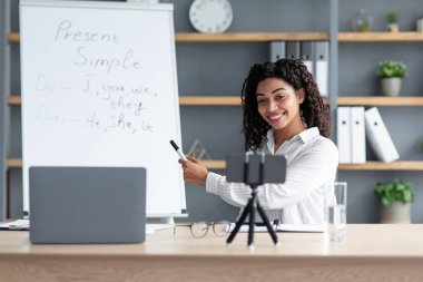 Eğitim, uzaklık çevrimiçi sınıf, görüntülü konuşma ve evde öğrenme. Neşeli bin yıllık Afro-Amerikan kadın öğretmen tahtaya işaret ediyor ve oturma odasındaki akıllı telefon web kamerasına bakıyor.
