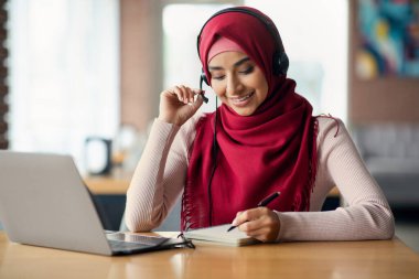 Orta Doğulu genç kız öğrenci online webinar 'a katılıyor, kafede masada oturuyor, dizüstü bilgisayar ve kulaklık kullanıyor, notlar alıyor, çevrimiçi dersler alıyor, hocaları dinliyor, boşluğu kopyalıyor.