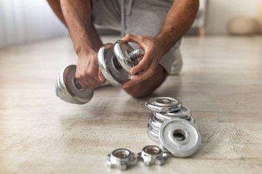 Ağırlık disklerini dambıllara takan genç adamın evde vücut geliştirme antrenmanına hazırlandığı yakın çekim görüntüsü. Tanımlanamayan bin yıllık adam içeride kas çalıştırıyor.