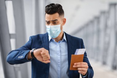 Tıbbi maskeli endişeli bir Arap kol saatine bakıyor, uçağa geç kalıyor, Orta Doğulu endişeli bir adam pasaport ve biletleri tutuyor, pandemik sırasında seyahat ediyor, yakın çekim sırasında kapısına doğru koşuyor,