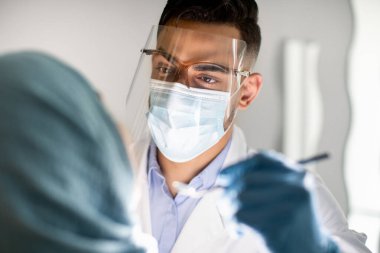 Arap Erkek Diş Hekimi Koruyucu Tıbbi Maske ve Yüz Koruyucusu Kadın Hastaların Dişlerini İnceleme, Stomatolog Doktor Diş Hekimliği ve Diş Hekimliği Kliniği 'nde