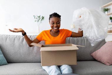 Memnun bir alıcı. Paketi alan heyecanlı siyahi kadın, karton paketleri açıyor, plastik ambalajı tutuyor, oturma odasında kanepede oturuyor. Genç Afro kadın internetten satın almaktan memnun.