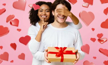 Siyah bir kadın elinde hediye tutarken erkek arkadaşının gözlerini şakayla kapatıyor. Çift gülümsüyor, kalplerle çevrili. Bu an bir kutlama sırasında neşe ve sevgi sergiliyor.