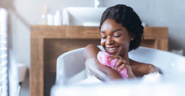 Bir kadın baloncuklarla dolu bir küvette oturuyor. Kolunu nazikçe pembe bir süngerle ovalıyor, mutlu bir şekilde gülümsüyor. Yakınlardaki bir pencereden banyoya ışık akıyor..