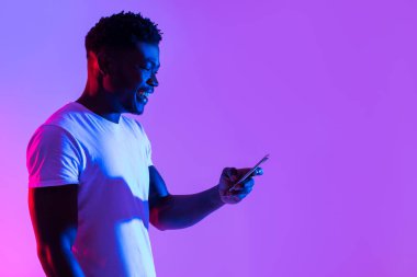 Neşeli genç siyah bir adam, sosyal medyada çalışmaya ya da paylaşmaya odaklanmış, neon ışıklı bir alanda akıllı telefonuyla meşgul oluyor. Modern teknolojiyi kullanarak yeni bir uygulamayı keşfederken heyecanını gösteriyor..