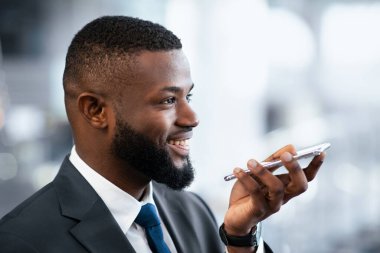 Sakallı bir adam resmi bir ortamda akıllı telefonuyla konuşuyor. Takım elbise giyiyor ve konuşurken gülümsüyor gibi görünüyor..