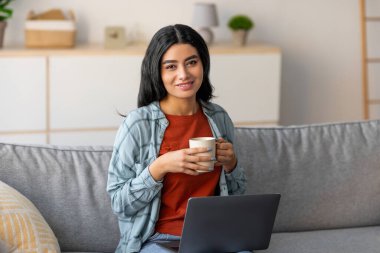 A beautiful young Arab woman sits comfortably at home, working on her laptop. She enjoys a cup of coffee while participating in an online meeting, showcasing remote work and learning.