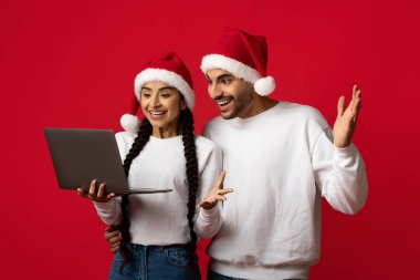Noel Baba şapkalı genç bir çift laptopta görüntülü sohbet ederken neşeli bir anı paylaşıyor. Parlak kırmızı arka planda heyecan ve mutluluğu ifade ederler. Bayram ruhu için mükemmeldir..