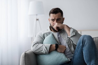 Olumsuz duyguların ifadesi, stres, depresyon, acı ve psikolojik sorunlar. Heyecanlı genç adam yastıkla kanepede oturur, yüzü elle kaplanır ve oturma odasında ağlar.