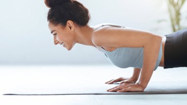 Bir kadın, kapalı bir alanda paspasın üzerinde şınav çekiyor. Gülümserken egzersizine odaklanıyor. Ortam enerjik ve fitness faaliyetlerini destekleyici görünüyor.